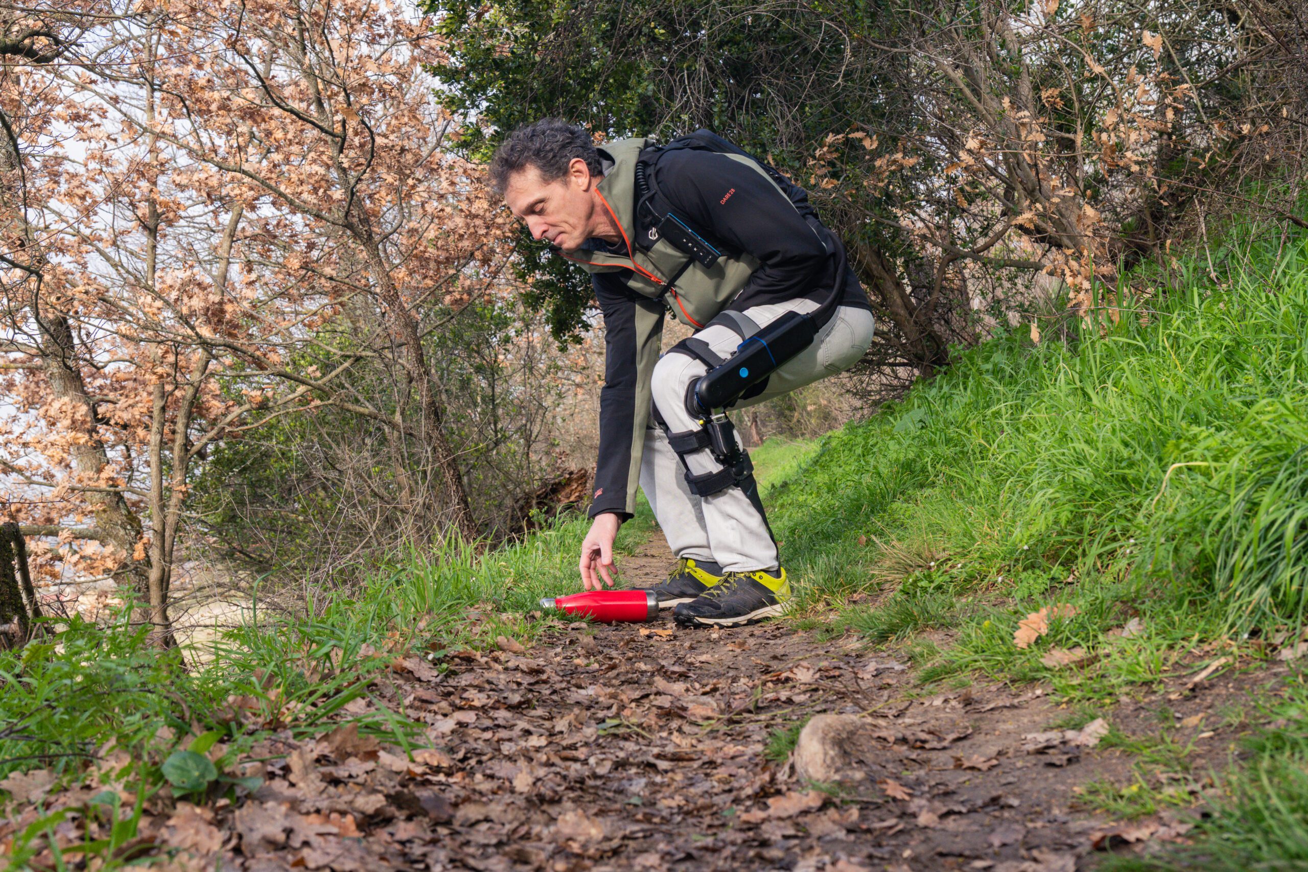 A man wearing a DREEVEN knee brace bends over to pick something up while walking on an outdoor hiking trail. 