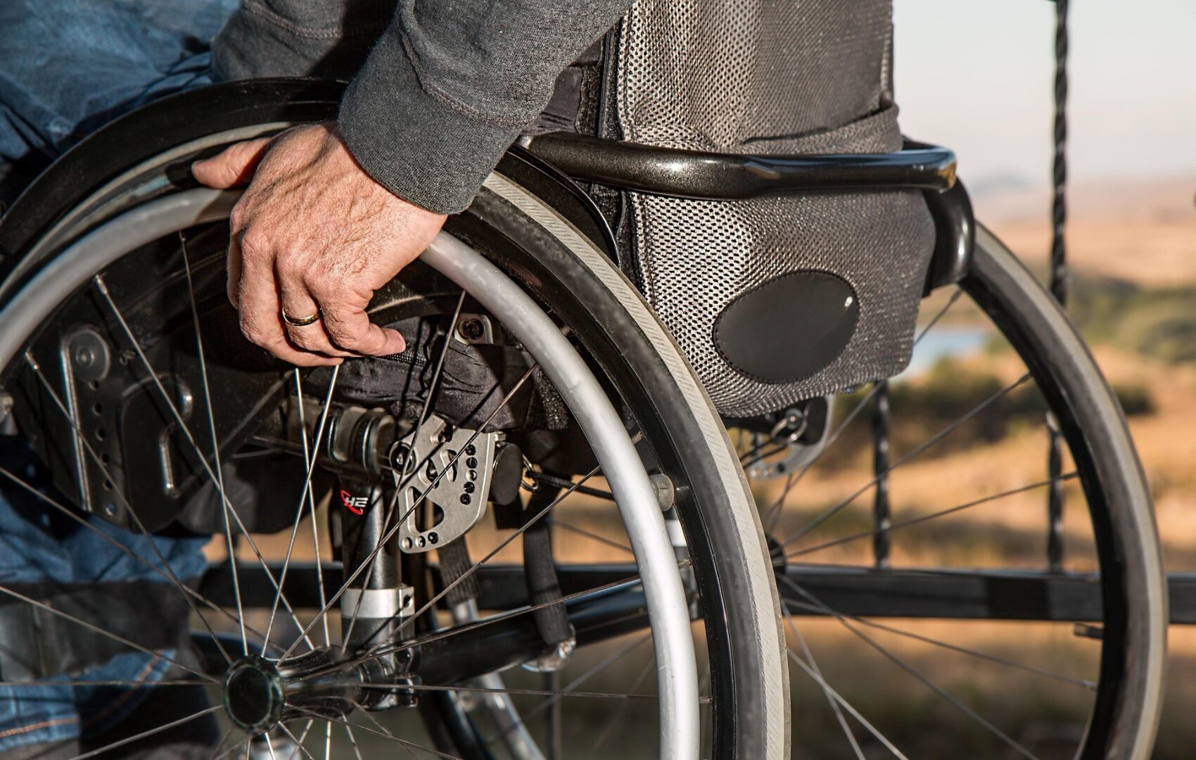 Upclose impage of a black/grey standard wheelchair from the side/behind. It shows an older man's hand resting on the push-bar, but doesn't show details of the rest of the person.